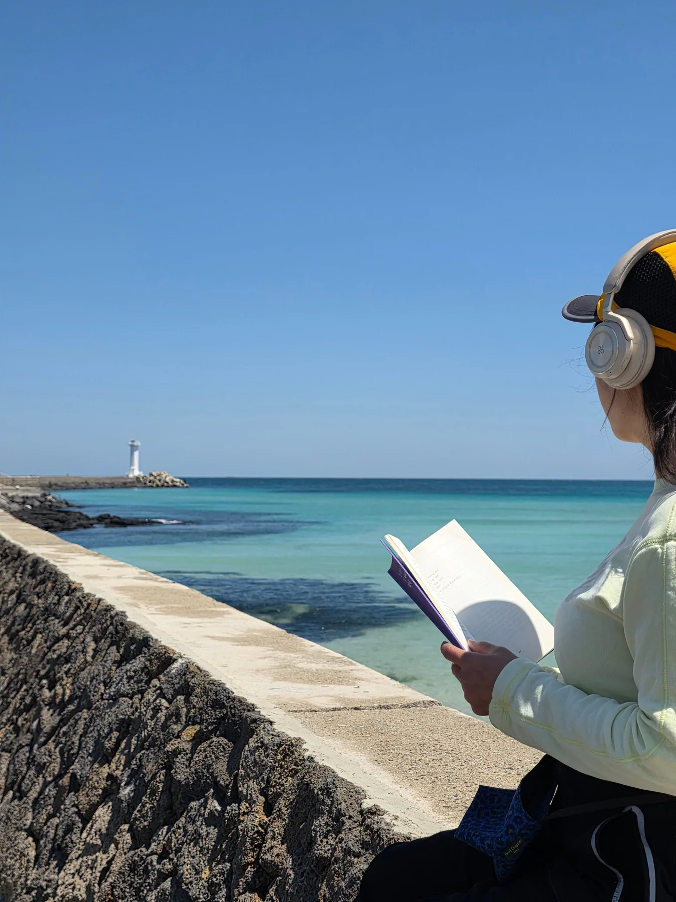 防波堤に座りヘッドホンをつけて本を読む旅人の後ろ姿、その先に翡翠色の海と白い灯台